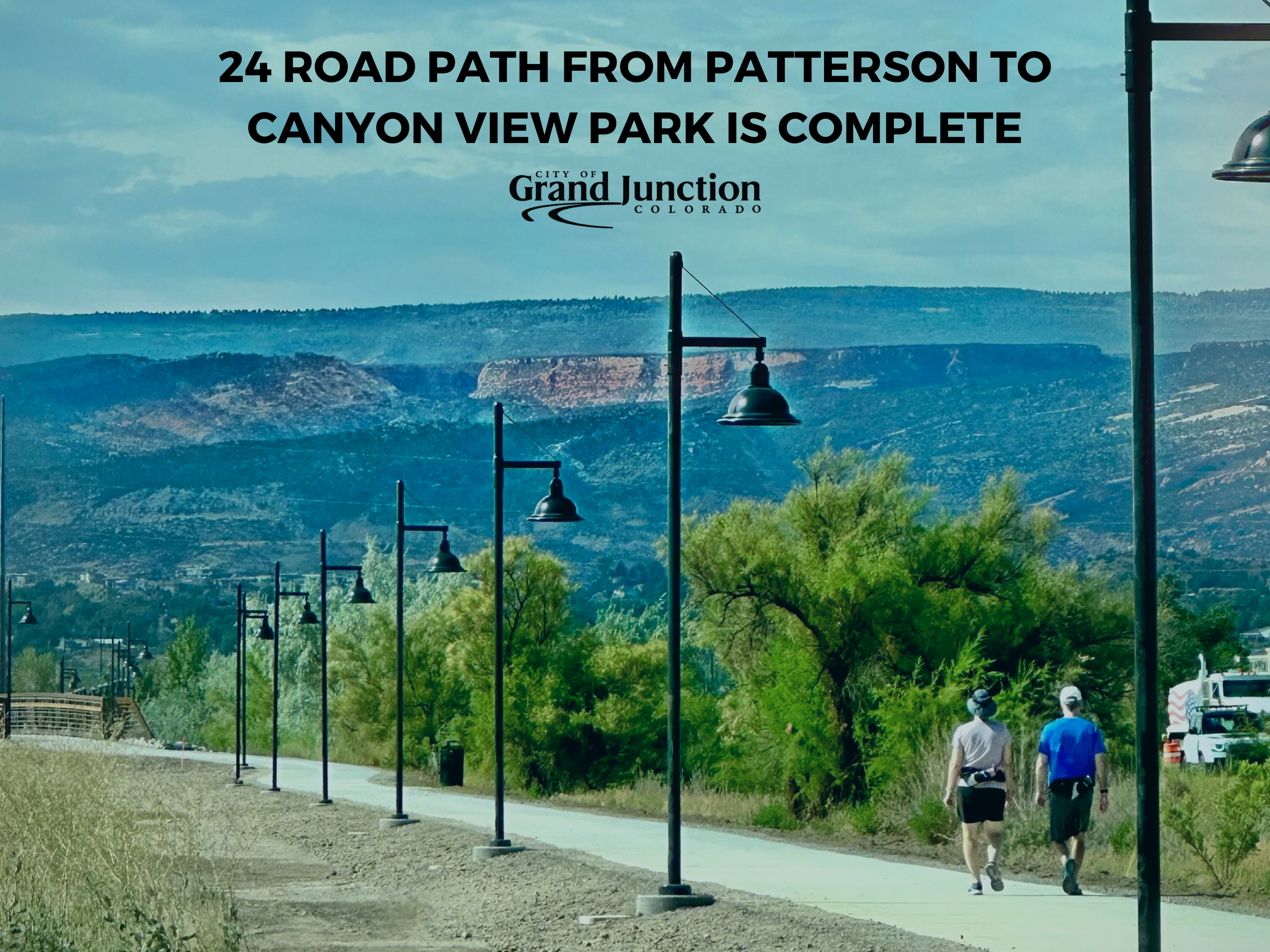 Two people walking along a path with light poles on the side. Mountains in the background and trees.